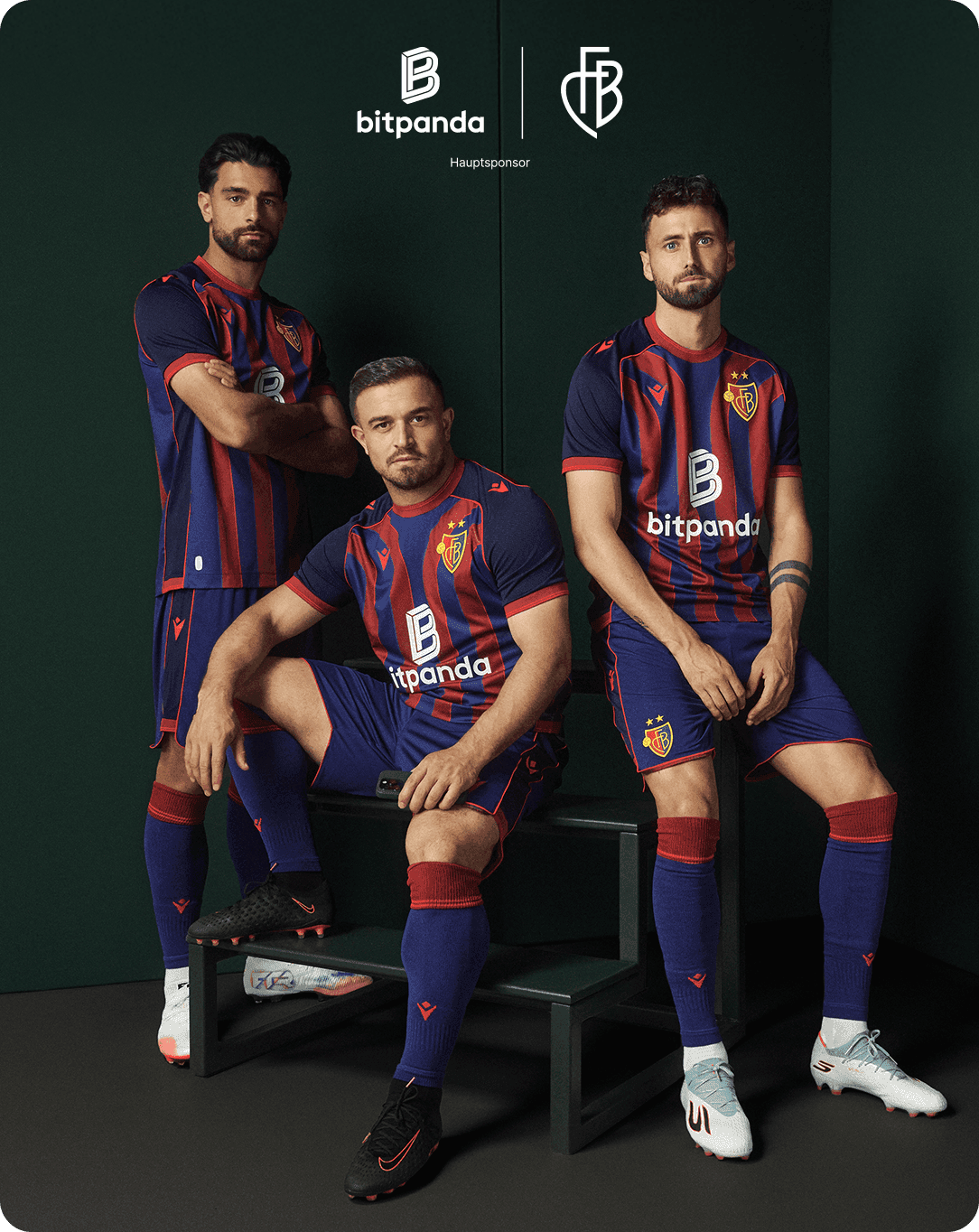 Three soccer players in red and blue striped uniforms pose confidently indoors with a green backdrop and sponsor logos visible.