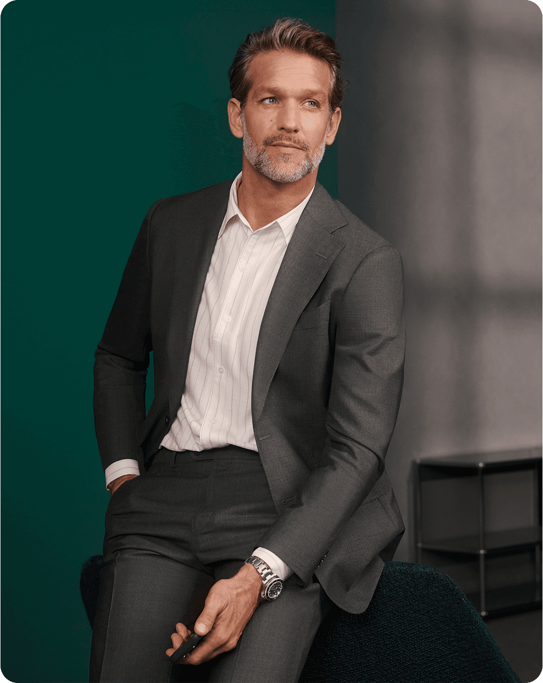 Man in a gray suit with a striped shirt, sitting and holding a phone, looking to the side, with a green background.