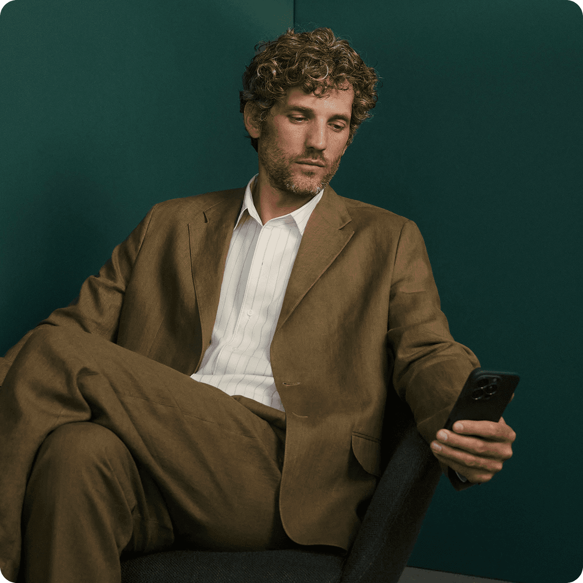 Man with curly hair in a brown suit sits on a chair, looking at his phone against a dark green background.