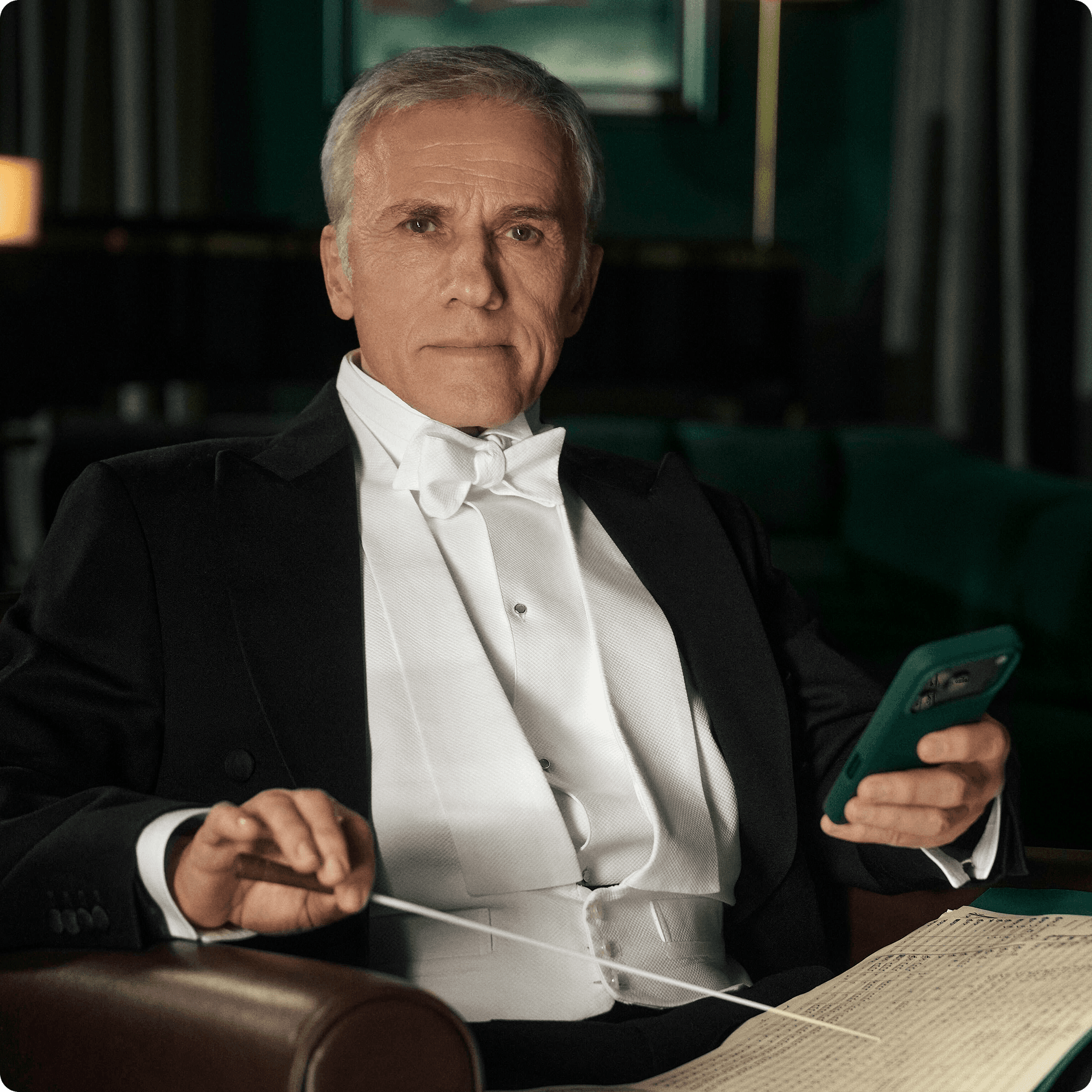 Christoph Waltz in a tuxedo sits in an armchair, holding a smartphone and conductor’s baton beside sheet music in a dim, elegant room.